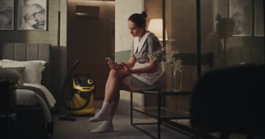 Yorgun Temizlik işçisi temizlik yaptıktan sonra nefes almak için ara veriyor. Profesyonel Üniformalı Hizmetçi Sessizce Otel Odasında Kol Sandalyesinde, Sakin İfadesiyle Telefonu Kaydırıyor. Öz Bakımı İşteyken kavra. El bilgisayarı.