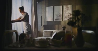 Stylish Hotel Suite 'de Üniformalı Kadın Temizlikçi Yatak ve Açık Perdeleri Düzeltti. Otel personeli konsept sabah rutini, özel hizmet, konfor, misafir deneyimi. Sabit Çekim.