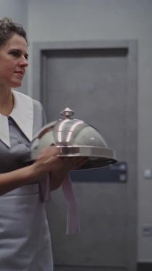 Gri üniformalı, Dost canlısı Bayan Otel Görevlisi Oda Servisi 'nden. Gümüş Kubbe şekilli süslemeli tepsi taşıyan bir kadın. Premium Catering ve Lüks Konukseverlik. Dikey Çekim.