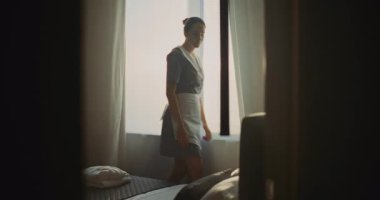 Bir sonraki konuğumuz için üniformalı bir otel görevlisi hazırlanıyor. Kadın perdeleri açar, yatağı düzeltir, bornozu katlar. Konsept Temizlik, Kalite Hizmeti ve Konforlu Seyahat Kalışı.