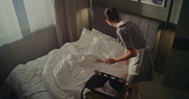 Vagonlu Otel Temizlik Personeli Yatak Çarşaflarını Değiştiriyor Konuk Gidiş. Kadın dikkatlice havluları kaldırır ve katlar ve yastıklardan yastık kılıflarını çıkarır. Odadaki Yüksek Temizlik Standartlarını Kabul Et.