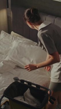 Vagonlu Otel Temizlik Personeli Yatak Çarşaflarını Değiştiriyor Konuk Gidiş. Kadın dikkatlice havluları kaldırır ve katlar ve yastıklardan yastık kılıflarını çıkarır. Odada Yüksek Temizlik Standartları. Dikey Çekim.