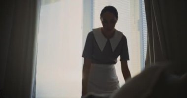 Parlak Misafir Odasında Kadın Otel Hizmetçisi Üniformalı Eldiven ve Temizlik Eldiveni, Temiz Yatak Çarşafları 'nda Gün Işığına İzin Vermek İçin Perdeleri Açıyor. Mesleki Konukseverlik Servisini Kabul Et.
