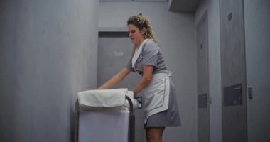 Otel Temizlikçisi Kadın, Temizlik Arabasının Yanında Duruyor, Temiz Havluları Pürüzsüz Katlıyor, Alıştırmalı Hareketler Yapıyor. Konukseverlik Profesyonelliği, Kalite Odası Hazırlığı ve Konuk Bakımı. Doli Vuruşu.