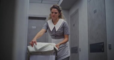 Temiz Üniformalı Kadın Otel Temizlikçisi, Temizlik Trolleyle Yanyana Duruyor, Taze Beyaz Havluları Güzelce Katlıyor. Konsept Temizlik, Düzen ve Yüksek Konukseverlik Hizmeti Standartları.