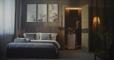 Üniformalı genç otel çalışanı lüks oteldeki modern odaya servis arabası getiriyor. Kadın Enfes Kahvaltı ve Taze Çiçekleri Odaya Getirdi. Konfor ve Premium Konukseverlik Deneyimi.