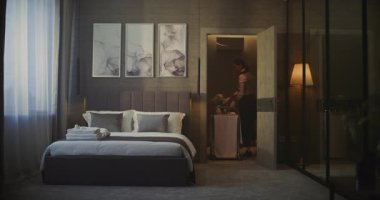 Otel hizmetçisi kahvaltı arabasıyla Zarif Otel Odasına giriyor. Şık Dekor 'la rahat bir iç mekan, Yatağın Yanındaki Kadın Kahvaltı Arabası. Konuk Bakımı, Profesyonel Otel Hizmeti Rutini. Sabit Çekim.