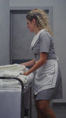 Profesyonel Üniformalı Kadın Koridorun aşağısındaki Tramvayı Temizliyor, Odaları Temizlemek için Temiz Havluları Taşıyor. Yüksek Kalite Otel Hizmetleri, Temizlik İşleri. Dikey Çekim.