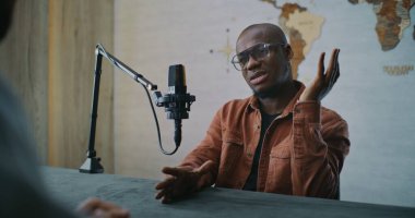 Podcast Konuğu Düşünceli Stüdyo Tartışması Yapıyor. Modern Podcast Stüdyosunda Afro-Amerikalı Adam Düşünceli Diyaloga Katılıyor, Karşılıklı Sunucu Masasına Karşılık Verirken Gülümsüyor. Portre.