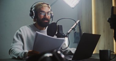 Profesyonel Erkek Podcast Sunucusu Stüdyoda Kayıt Bölümü. Gözlük ve kulaklık takan Sakallı Adam Ses Stüdyosunda Senaryodan Mikrofona Okuyor, Prodüksiyon Dijital Medya İçeriğine Odaklanıyor.