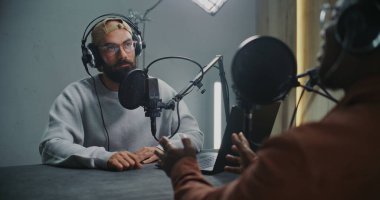 Kulaklık takan ve Profesyonel Mikrofonlara Konuşan İki Adam, Ciddi Ton 'u korurken fikir alışverişinde bulundular. Modern Stüdyo Kurulumunda Bilgisayar Kayıt Podcast 'i Olarak Sunucu ve Misafir Arasında Oturur.