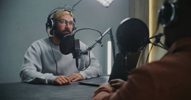 Kulaklık takan ve Profesyonel Mikrofonlara Konuşan İki Adam, Ciddi Ton 'u korurken fikir alışverişinde bulundular. Modern Stüdyo Kurulumunda Bilgisayar Kayıt Podcast 'i Olarak Sunucu ve Misafir Arasında Oturur.