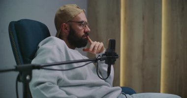 Sakallı Podcast sunucusu Kaptan ve Gözlük Hareketleri Profesyonel Stüdyo Mikrofonuna Konuşurken. Yetişkin Adam Dinamik Kayıt Oturumunda Arkaya Yaslanıyor, Görüşlerini Paylaşıyor.