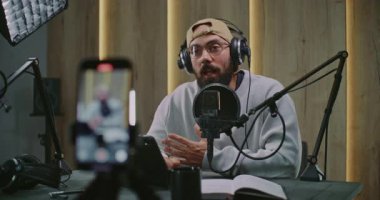 Akıllı Telefon Kayıtları Sakallı Podcast Sunucusu Masanın Ötesinde Konuşuyor, Misafir Hareketlerine Yanıt Veriyor. Cup, Phone ve Yetişkin Adam 'ın yanındaki masa üzerine kitap. Otantik Stüdyo Ayarları Dikey Çekimde Yakalandı.