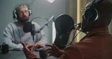 İki Podcast sunucusu Laptop, Pro Mikrofonlar ve Sıcak İçecekler Masanın karşısında Aktif Sohbet Ettiler. Erkekler fikir alışverişinde bulunurlar, birbirlerini işaret ederler ve doğal olarak tartışmayı sürdürürler..