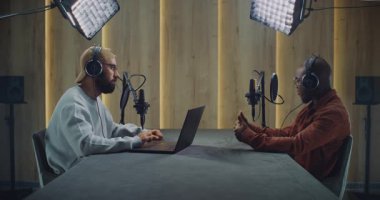 Sakallı Podcast sunucusu, Afro-Amerikan konuğu ile röportaj yaparken dizüstü bilgisayar kullanıyor. Gerek kulaklıkla, gerekse stüdyodaki mikrofonlarla konuşun ve odaklanmış konuşma sırasında doğal olarak hareket edin..