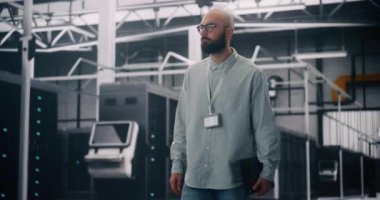 Modern Veri Merkezi 'nde Bilgi İşlem Mühendisi Sunucu Raflarında Yürüyor. Rozetli Odaklanmış Adam ve Parlayan Sunucu Rafları Arasında Dizüstü Bilgisayar Dolaşıyor. Bilişim Altyapısı ve Bulut Teknolojisi kavramı.