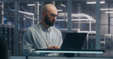 Sunucu Salonunda dizüstü bilgisayar kullanan odaklı bilişim mühendisi. Açık Gömlekli Adam Duruyor ve Gizlice Çalışıyor, Arka plan Sunucu Modern Veri Merkezi 'ni Gösteriyor. Alan Teşhisi ve Sistem Bakımı Konsepti.