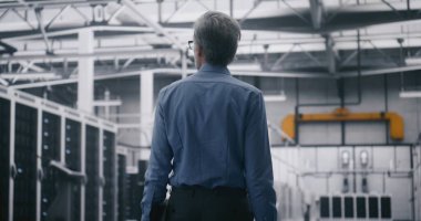 Yetkili Denetleme Merkezi Akışı. Gri Saçlı Adam devasa ölçekli veri merkezinde dizüstü bilgisayar taşıyor. Stratejik Gözetim ve Teknolojik Altyapı Yönetimi. Arka Görünüm.