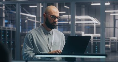 Sunucu Salonunda dizüstü bilgisayar kullanan odaklı bilişim mühendisi. Açık Gömlekli Adam Duruyor ve Gizlice Çalışıyor, Arka plan Sunucu Modern Veri Merkezi 'ni Gösteriyor. Alan Teşhisi ve Sistem Bakımı Konsepti.