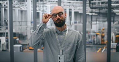 Gözlüklü sakallı mühendis, profesyonel tavır takınıyor. Modern İmalat Tesisinin Yüksek Teknoloji Ortamı 'nda Ayakta Duran Adam. Endüstride Teknik Uzmanlık ve Yenilik 4.0.