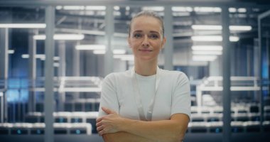 Kolları çaprazlanmış gülümseyen kendinden emin kadın bilişim lideri. Modern Veri Merkezi 'nde, Güvenliğin Ortasında Duran Genç Kadın. Profesyonellik, Teknoloji Uzmanlığı ve Otorite kavramı. Portre.