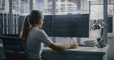 Kadın Teknik Uzman Veri Merkezi 'ndeki Çoklu Ekran Kurulumunda Ölçüm ve Grafik Verilerini İzlerken Kulaklık Aracılığıyla İletişim Kuruyor. Kontrol ve Destek, Uzman Danışmanlık ve Karmaşık Problem Çözme.