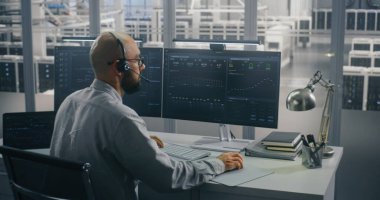 Uzman Koordinasyon Veri Merkezi Operasyonları Uzaktan. Kulaklık üzerine konuşan bilişim uzmanı, Yüksek Güvenlik Veri Merkezi 'ndeki çift monitörlü masada aktif olarak yazıyor. Odaklanmış Veri Analizi Konsepti.