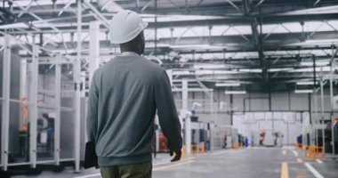 Sanayi Denetleyicisi Otomatik Üretim Sürecini Robotik Fabrika Katında İzliyor. Koruyucu Sert Şapka, Güvenlik Uyumluluğu ve Kompleks Tesisinin Profesyonel Yönetimini Vurguluyor. Arka Görünüm.