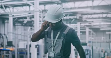 Endüstriyel Erkek Mühendis, Smartphone 'da konuşurken ve gülümserken Yüksek Teknoloji Fabrikası' nın zemininde yürüyor ve meslektaşıyla tartışıyor. Akıllı Üretim, Uzaktan Koordinasyon ve Endüstri 4.0 Konseptleri.