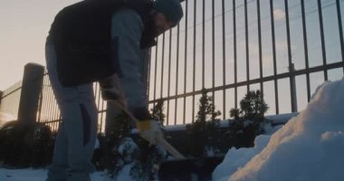 Kış için Giyinen Erkek, Evin girişinde kar temizliyor. Örgü Beanie 'li Adam ve İş Giysisi Yeleği Kar Kaldırma Üzerine Odaklanıyor. Günlük Ev İşleri, Kış Koşullarında Sorumlu Mülkiyet Bakımı.