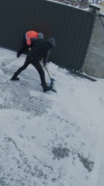 Erkekler garaj kapısının yanındaki kürekli karları temizler. Turuncu Ceketli İşçi ve Siyah Yelekli Adam Paved Arka Bahçesinde İşbirliği Yapıyor. Güvenlik kamerası Kış Malları Bakımı 'nı gösteriyor. Dikey Çekim.