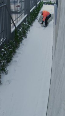 Yüksek Görüş Alanı Ceketli İşçi Dar Kaldırımdan Kar ve Buz Temizliyor. Kış Bakım Faaliyeti Dış Güvenlik Kamerası 'nı kaydetti. Kamu Güvenliği, Soğuk Hava İşleri. Dikey Çekim.