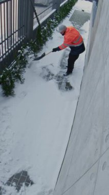 Yansıtıcı Ceket Giyen Erkek Kamu Hizmetleri İşçisi Kış Koşullarında Çitin Yanı Başındaki Çitten Karı Kaldırdı. Güvenli yaya girişi, mevsimlik bakım, gözetleme kamerası. Dikey Çekim.