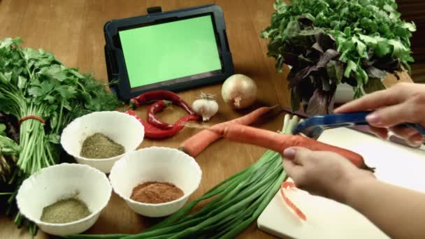 La mujer limpia la zanahoria y observa la receta en la tableta — Vídeo de stock