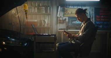 Profesyonel Garaj 'da, Erkek Teknisyen Sandalyede Oturuyor, Tablet' te Gerçek Zaman Verilerini Analiz Etmeye Çalışıyor. Mobil Çalışma İstasyonundaki Dizüstü bilgisayar Kompleks Tanı Grafikleri ve Araba Şemaları 'nı da gösterir.