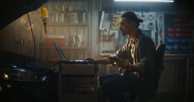 Profesyonel Garaj 'da, Erkek Teknisyen Sandalyede Oturuyor, Tablet' te Gerçek Zaman Verilerini Analiz Etmeye Çalışıyor. Mobil Çalışma İstasyonundaki Dizüstü bilgisayar Kompleks Tanı Grafikleri ve Araba Şemaları 'nı da gösterir.