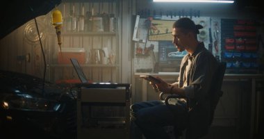 Profesyonel Garaj 'da, Erkek Teknisyen Sandalyede Oturuyor, Tablet' te Gerçek Zaman Verilerini Analiz Etmeye Çalışıyor. Mobil Çalışma İstasyonundaki Dizüstü bilgisayar Kompleks Tanı Grafikleri ve Araba Şemaları 'nı da gösterir.