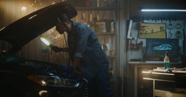 Adam iyi donatılmış bir garajda kapüşonlu açık bir şekilde duruyor. Parlak hizmet lambası altında teknik denetim yapıyor. Guy Motor Bileşenlerini İnceledi, Uygun Bakım, Araç Performansı Sağlama.