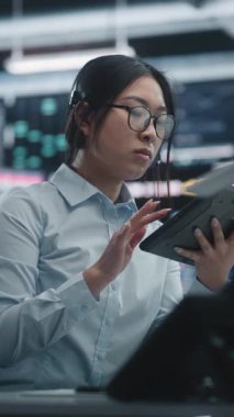 Tablet ve Çoklu İzleme Alanıyla Asya Kadın Analiz Eğilimleri. Gözlüklü Kadın İş İstasyonunda Oturuyor, Dijital Tablet 'teki Gerçek Zaman Verilerini Çapraz Referans Veriyor ve Kulaklıkla Konuşuyor. Dikey Çekim.