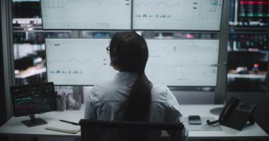 Sofistike İş İstasyonunda çalışan kulaklıklı bir tüccar. Karmaşık borsa grafikleri ve gerçek zamanlı analizlerle dolu dört büyük ekranı izlerken kadın dijital tabletle etkileşime giriyor. Arka Görünüm.