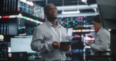 Üst Düzey Komisyoncu, piyasayı sakin bir şekilde analiz ediyor. Siyah Erkek Kulaklık ve İp Takan, Yüksek Teknoloji Borsasında Duran, Dijital Tableti Tutan ve Veri Ekranlarına Düşünerek Bakan.