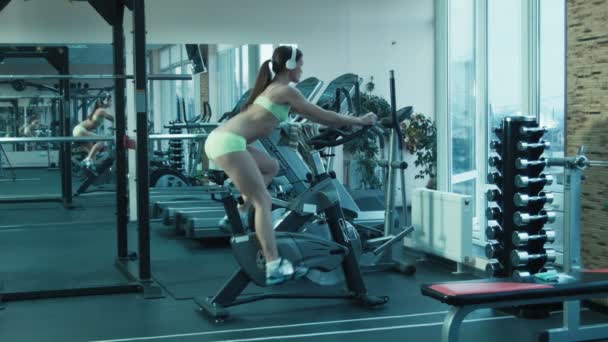 Спортсменка з навушниками, що розмовляють на симуляторі на стаціонарному велосипеді у спортзалі . — стокове відео