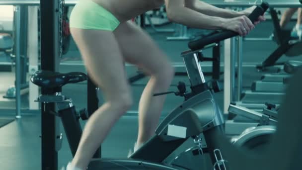 Mujer atlética con auriculares pedaleando en el simulador en una bicicleta estacionaria en el gimnasio . — Vídeos de Stock