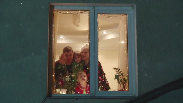 La famille heureuse regarde la neige à travers une fenêtre et salue les mains — Video