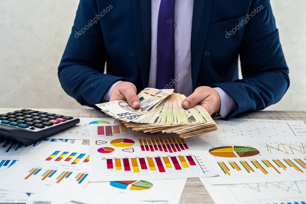young businessman in office counts money as profit after a successful deal. The man compares and points to profit. Business graphs, Ukrainian hryvnia money on the table.