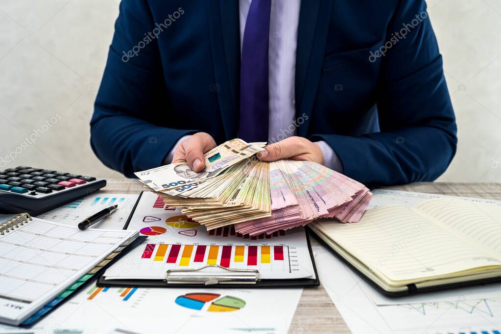 young businessman in office counts money as profit after a successful deal. The man compares and points to profit. Business graphs, Ukrainian hryvnia money on the table.
