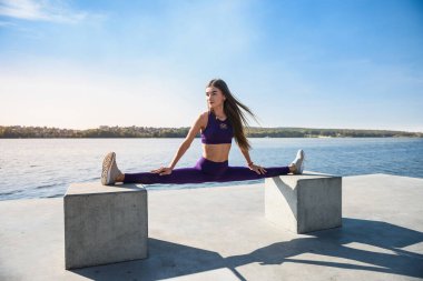 Genç Avrupalı bayan sporcu bacaklarını esnetme egzersizleri yapıyor, dışarıda göl kenarındaki yarıklara oturuyor. Sağlıklı yaşam tarzı