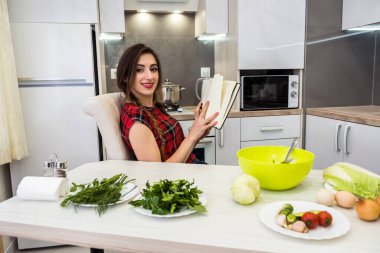 Genç ve güzel bir kadın mutfak masasında farklı sebzelerle oturuyor ve günlüğüne sağlıklı beslenmeyle ilgili her şeyi yazıyor ya da yazıyor.