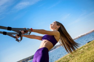 Spor elbiseli güzel bir kadın gündüz vakti trx fitness kayışlarıyla göl kenarında şınav çekiyor. Sağlıklı yaşam tarzı